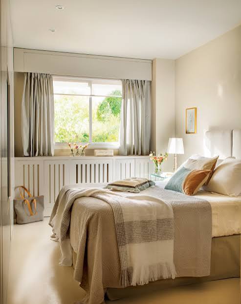 Dormitorio luminoso con una cama, ventanas grandes y decoraciones suaves en tonos neutros.