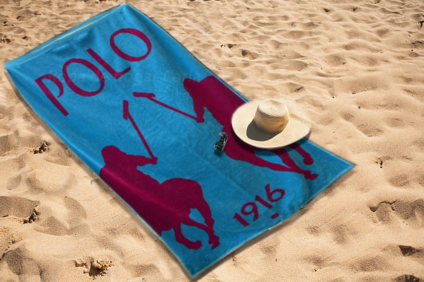 Toalla de playa azul con diseño de polo y un sombrero sobre la arena.