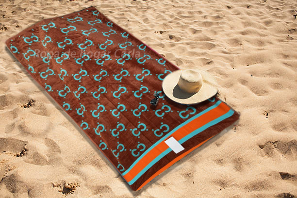 Toalla de playa marrón con patrón, junto a un sombrero, sobre arena clara.