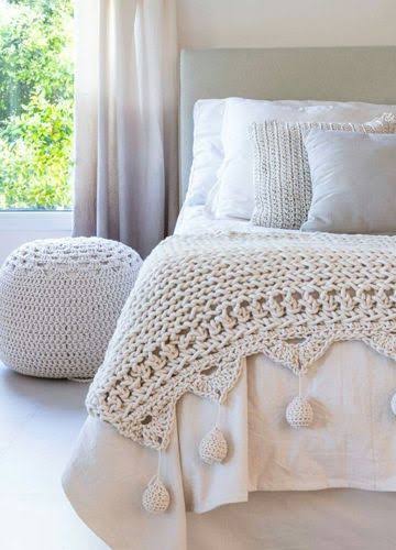Cama con manta de ganchillo blanca y cojines, junto a una mesa redonda blanca.
