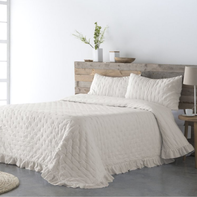 Cama con edredón claro y almohadas, entorno minimalista y decoración en tonos neutros.