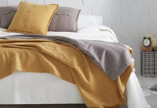 Cama con sábanas blancas, manta gris y cobertor amarillo, decorada con almohadas.