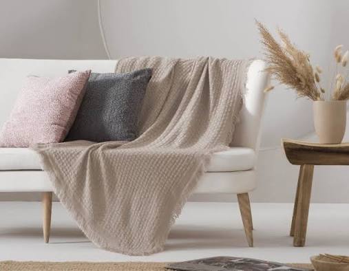 Sofá blanco con cojines grises y rosados, y una manta beige sobre él. Decoración minimalista.