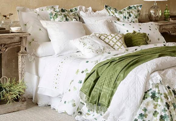 Cama con sábanas blancas y cojines verdes y florales, decorada con un ambiente acogedor.