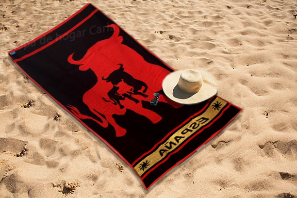 Toalla de playa negra con diseño de toro rojo y sombrero de paja al lado en la arena.