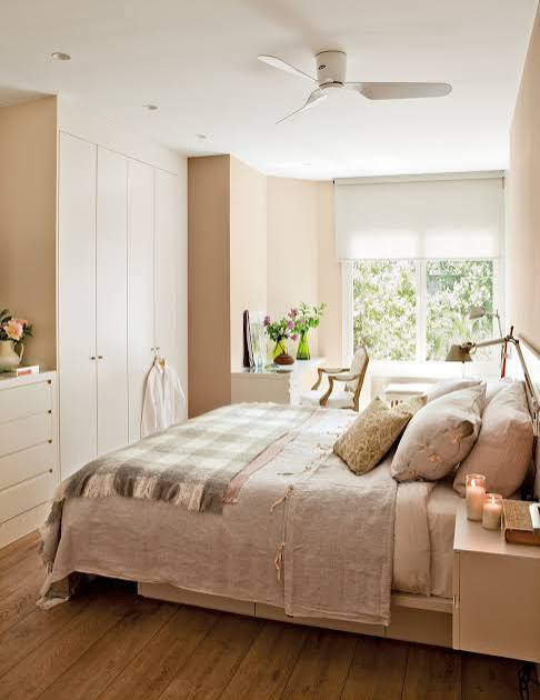 Habitación luminosa con cama, armario blanco y ventanales, decorada con flores.