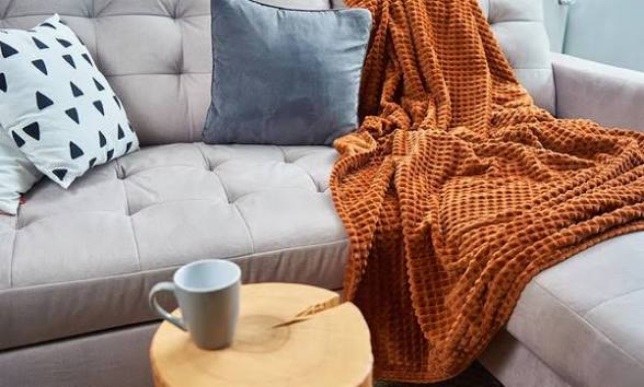 Sofá con manta marrón, cojines decorativos y taza en mesita de madera.