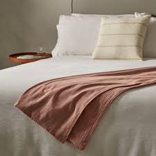 Manta de color rosado sobre una cama con almohadas blancas y una mesita de noche.